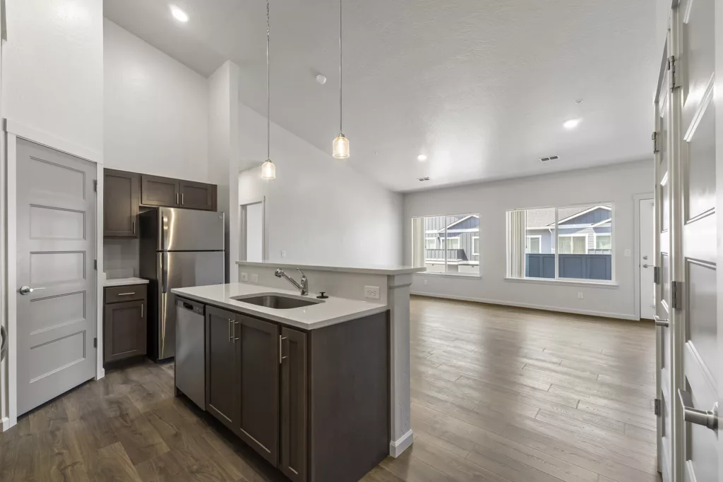 A modern kitchen and living area featuring wooden flooring, an island with a sink, dark brown cabinets, stainless steel appliances, and large windows allowing natural light to fill the space.