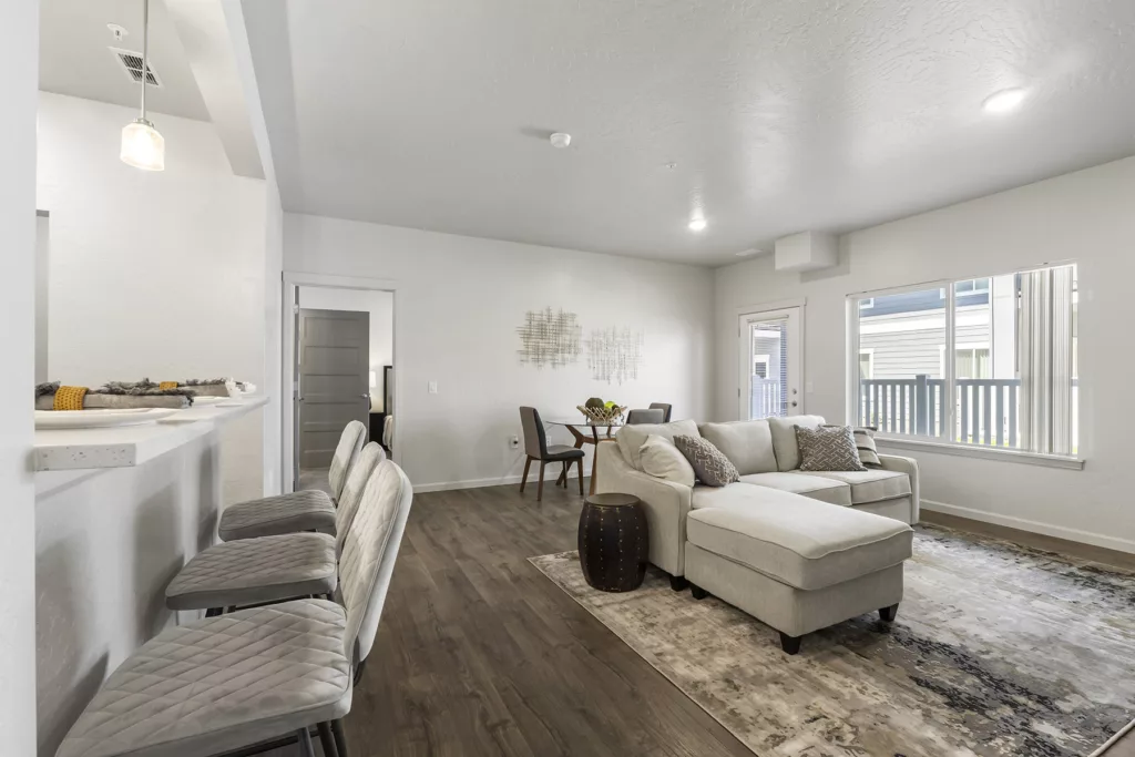 A modern living room with wooden flooring, featuring a light gray sectional sofa, a textured rug, and a small round coffee table. The room has a dining area with a round table and chairs in the background, and large windows on the right side provide natural light. A raised countertop with bar stools is visible on the left.