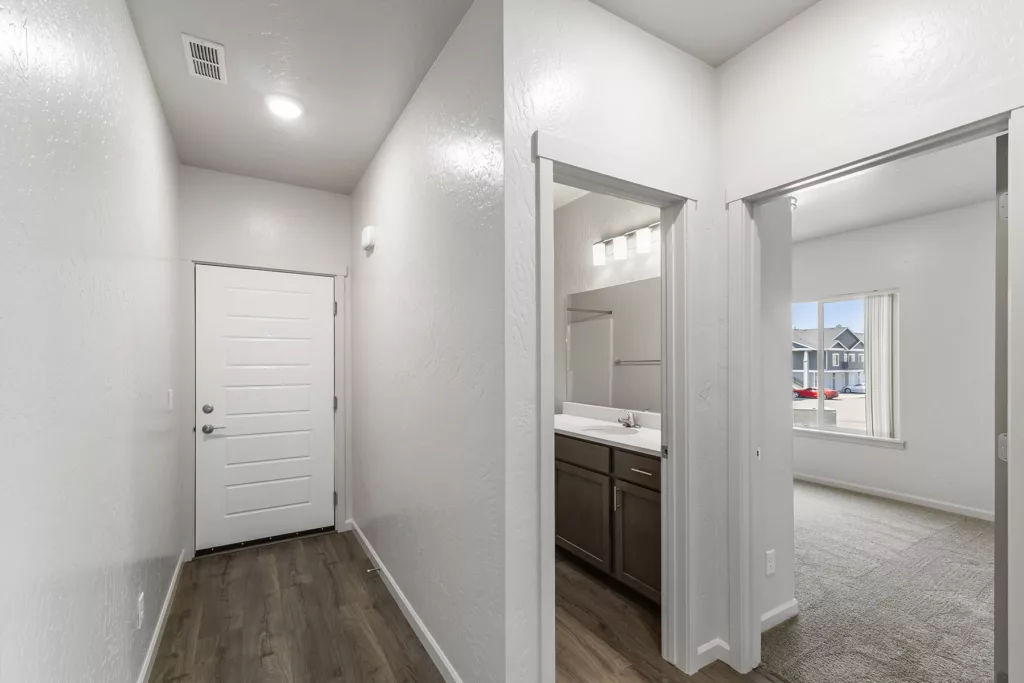 A modern home interior showing a hallway with wooden flooring, leading to a white door. On the right, there is a glimpse of a bathroom with a sink, mirror, and cabinets, and a room with a window looking out to a street with cars and houses.
