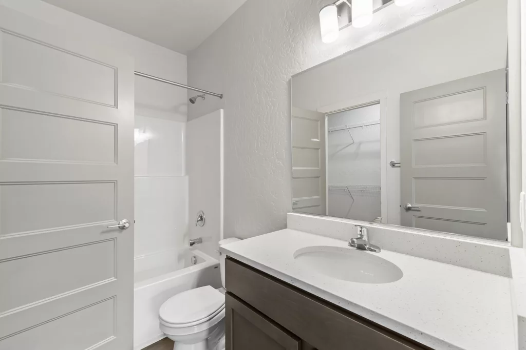 A modern bathroom featuring a white countertop with an oval sink, a wooden cabinet, a toilet, a bathtub with a showerhead, and a large mirror. The room is well-lit with ceiling lights and has light gray walls and doors.