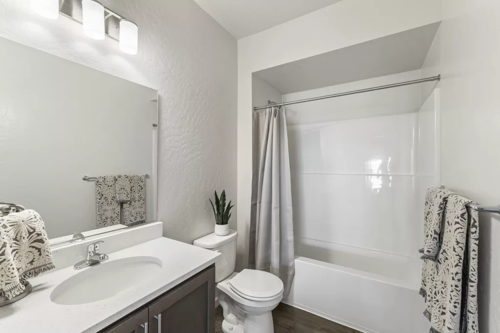 A modern bathroom with a white countertop and sink, a large mirror, and wooden cabinets. A toilet and a potted plant are positioned beside a shower-tub combination with a gray shower curtain. Towels with a decorative pattern hang on the towel rack and countertop. The room is well-lit with wall-mounted lights above the mirror.