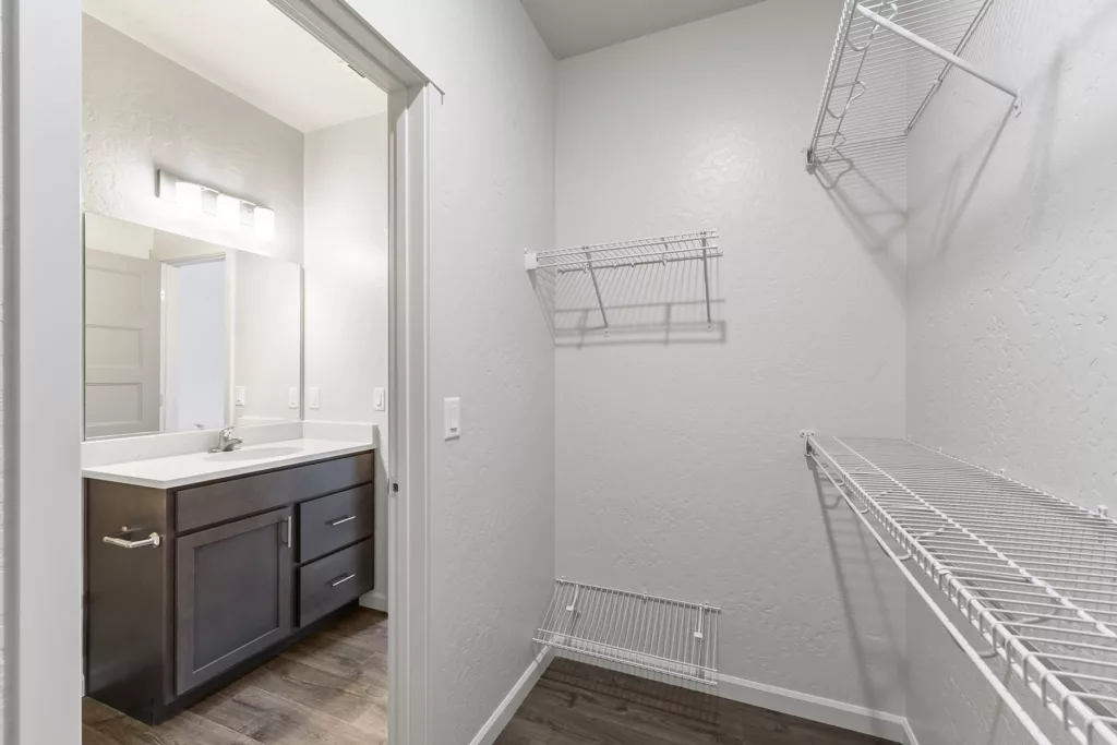 A walk-in closet with white wire shelving and a view into a bathroom featuring a dark wood vanity and sink, set against a light-colored wall and flooring.