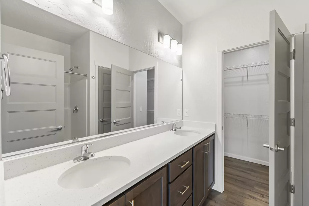 A modern bathroom featuring a double-sink vanity with a large mirror above it, a closed shower area on the left, and an open closet with wire shelving on the right. The space is well-lit with overhead lighting, and the overall color scheme is white and gray with wooden cabinetry.