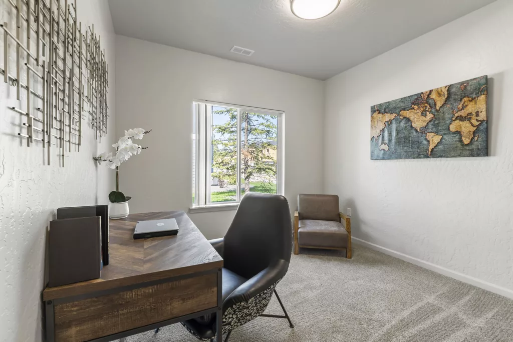 A small home office with a wooden desk and a black chair. A laptop is placed on the desk along with a white potted orchid. On the right wall, there's a map artwork, and a gray armchair is positioned nearby. A window lets in natural light, illuminating the carpeted floor and metal wall art on the left.