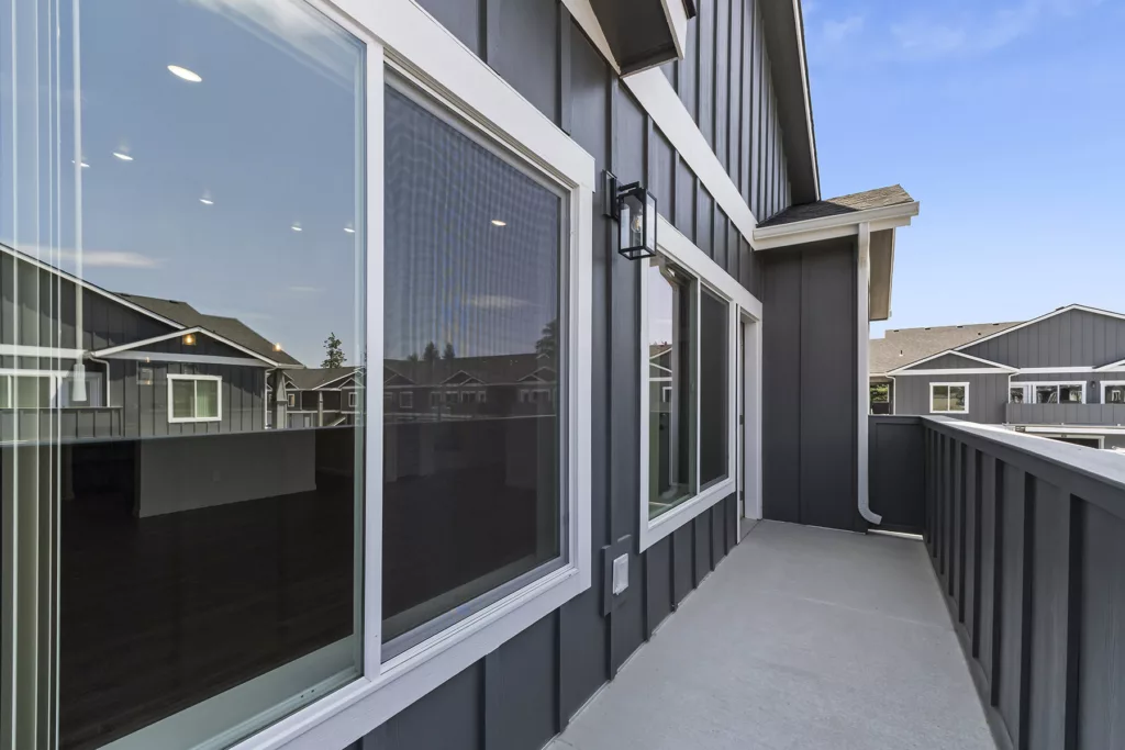 A modern balcony with dark gray siding and large windows reflecting nearby buildings, featuring a simple light fixture and a wooden railing under a clear blue sky.