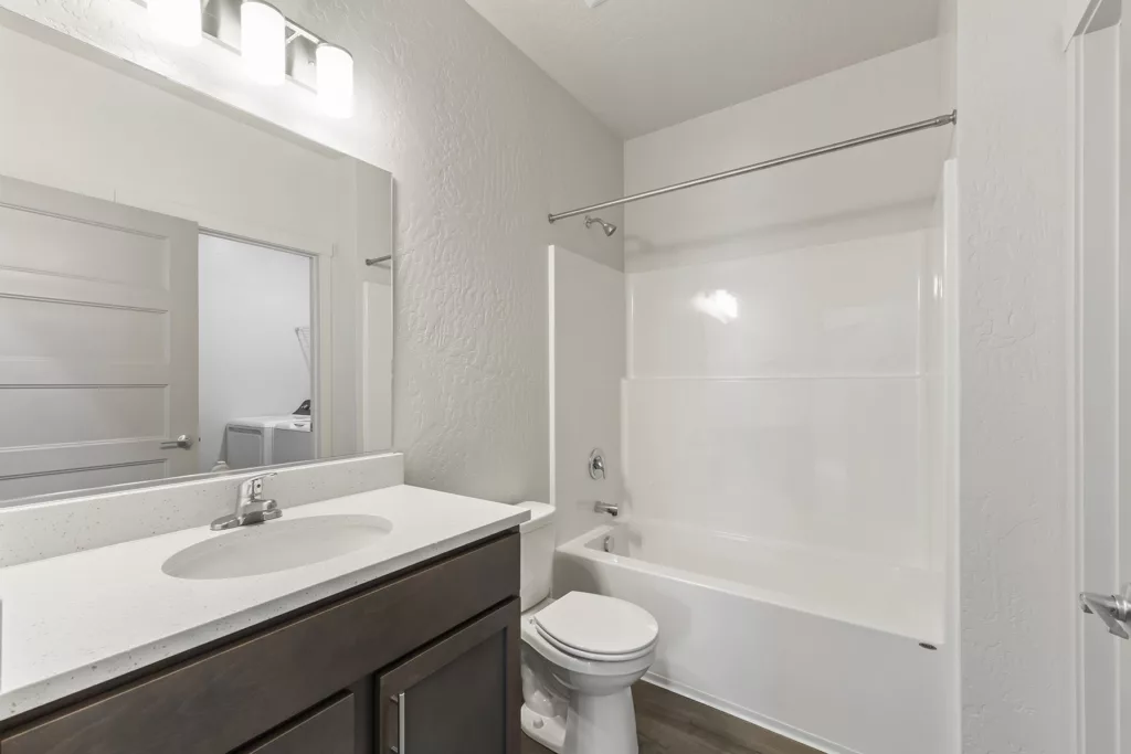 A modern bathroom with a white countertop sink, dark wooden vanity, toilet, and a bathtub with a showerhead. The room has neutral-colored walls and is well-lit by a fixture above the large mirror.