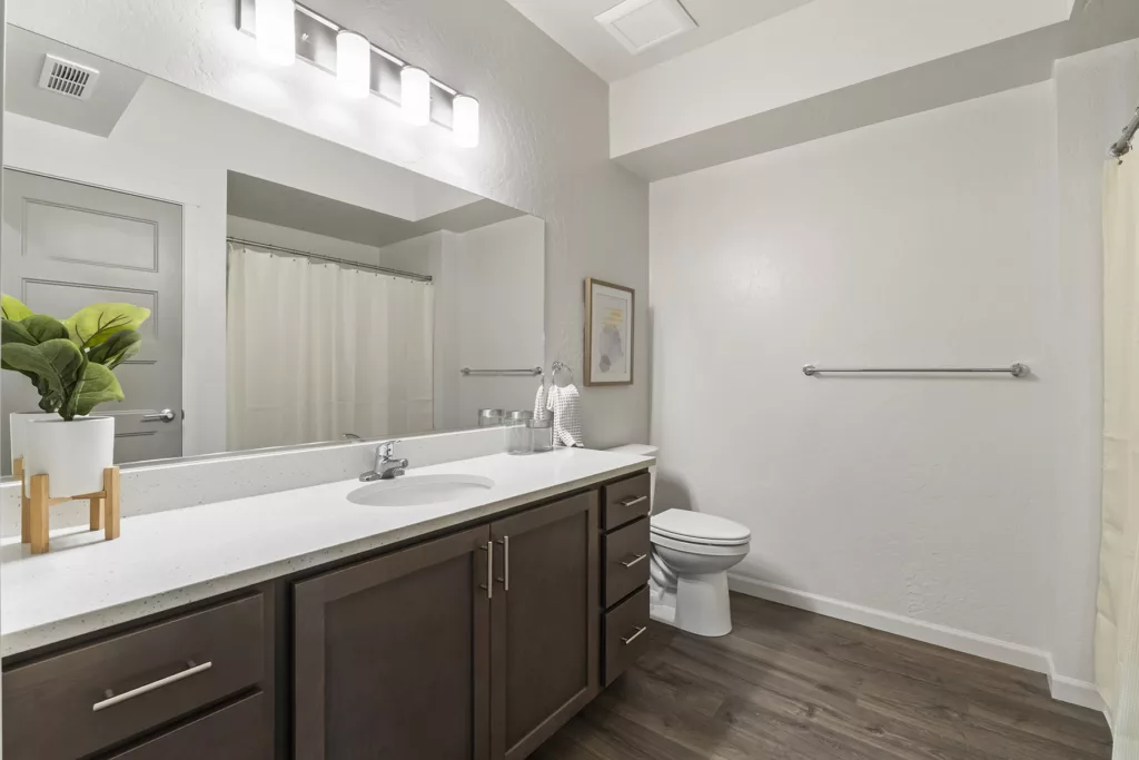 A modern bathroom with a large mirror above a dark wood vanity, featuring a single faucet, potted plant, and towels. The room includes a toilet, light fixture, and a shower with a cream-colored curtain, all set against white walls and a wood floor.