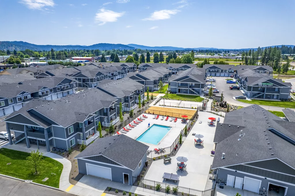 Aerial view of a residential complex with multiple gray-colored apartment buildings surrounding a central outdoor swimming pool with red lounge chairs and umbrellas. Mountains and greenery are visible in the distant background.