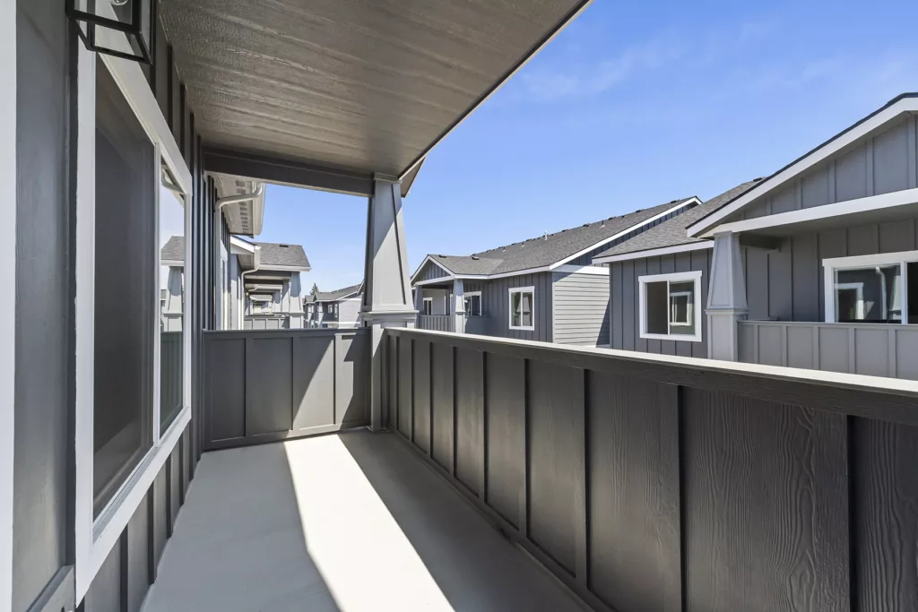 A view from a balcony with wooden railing and nearby houses with gray siding and white trim under a clear blue sky.