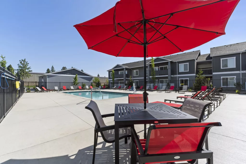 Outdoor swimming pool area with red lounge chairs and a table set under a bright red umbrella, in front of modern apartment buildings.