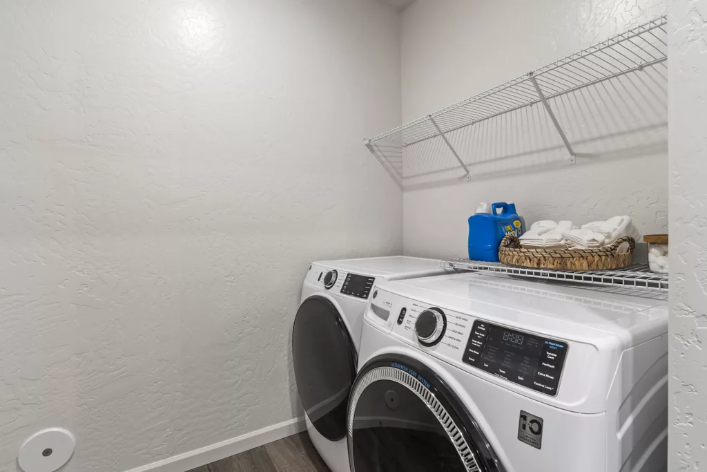A modern laundry room with a white front-loading washer and dryer side by side. Above them is a metal wire shelf holding laundry detergent, a basket of folded towels, and a jar of dryer sheets.