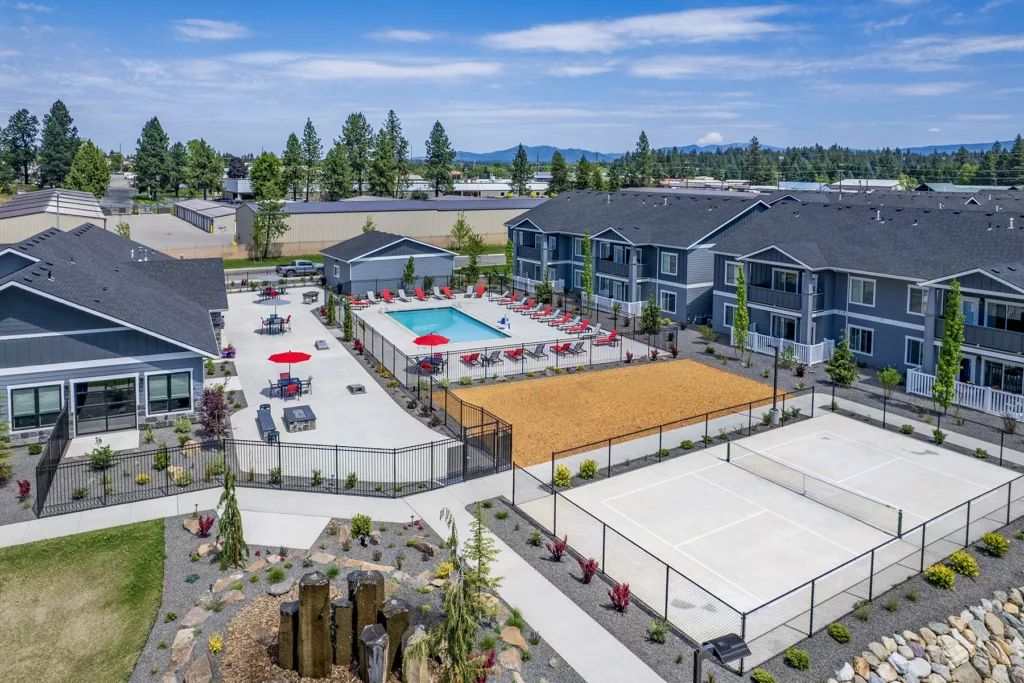Aerial view of a modern apartment complex featuring a swimming pool with red lounge chairs, adjacent to a tennis court and a bocce ball court, surrounded by landscaped gardens and pathways.
