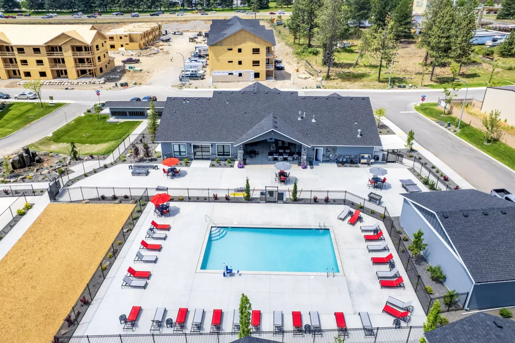 Aerial view of a modern residential complex featuring a swimming pool surrounded by red loungers and umbrellas. The well-maintained facility includes a clubhouse and landscaped areas, with construction visible in the background.