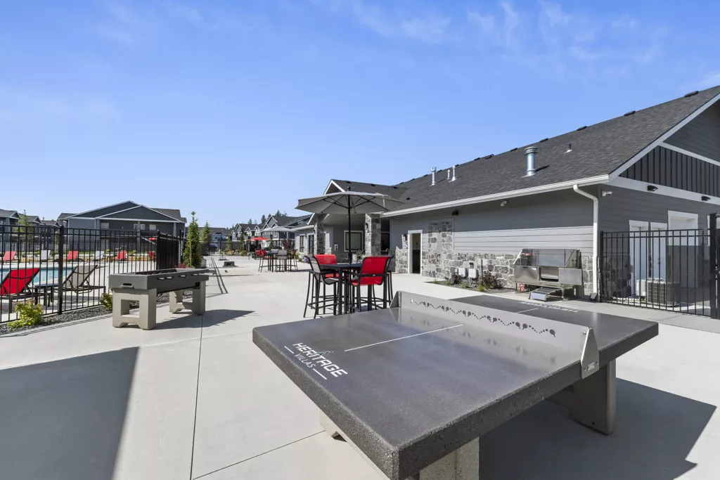 Outdoor recreation area with a ping pong table, foosball table, and seating under umbrellas, near a pool and modern buildings.