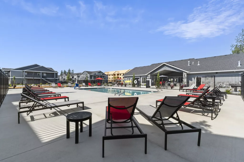 A modern outdoor pool area with several red and black lounge chairs arranged neatly around it, surrounded by a black metal fence. The background features a building complex with gray rooftops under a clear blue sky.