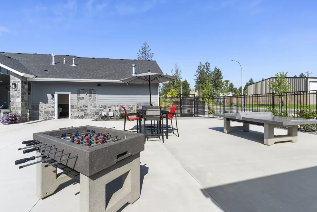 Outdoor recreational area with a foosball table and ping pong table under a clear blue sky, surrounded by a stone building and metal fence.