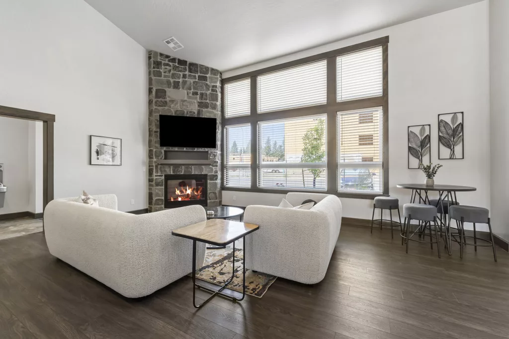 A modern living room with a stone fireplace and a wall-mounted TV above it. Two white sofas face the fireplace, with a wooden coffee table in the middle. Large windows provide natural light, and a small round table with stools is set against the wall.