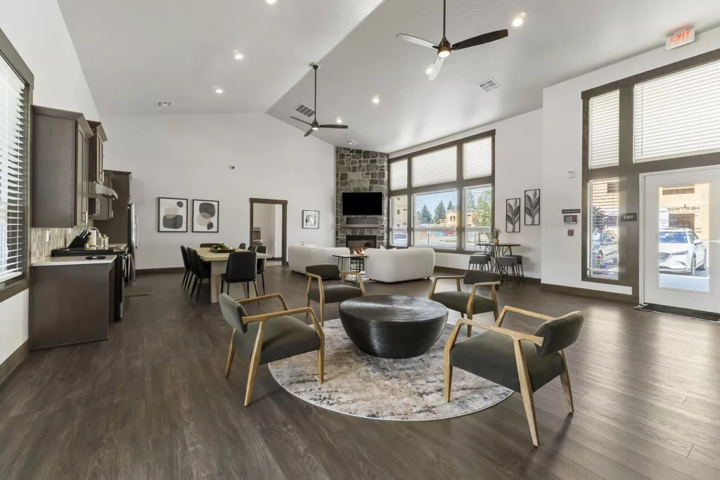 A spacious communal living area with modern decor, featuring a large round coffee table surrounded by four wooden armchairs with black cushions. The space includes a kitchen area with a dining table and chairs on the left, a stone fireplace and wall-mounted TV in the background, and large windows providing natural light. The room is lit by ceiling lights and fans, and decorative art hangs on the walls.