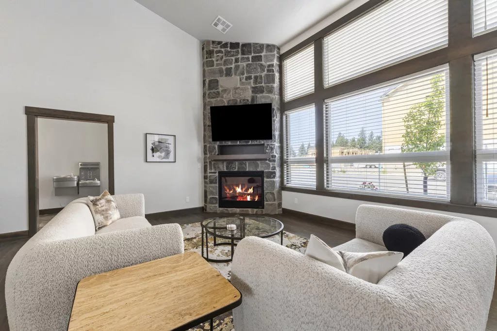 Modern living room with two white cushioned chairs facing a glass coffee table, a stone fireplace with a TV mounted above, and large windows with blinds on the right.