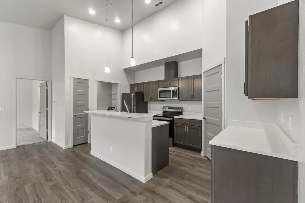 Modern kitchen with dark wood cabinets, white countertops, stainless steel appliances, and pendant lighting, featuring a high ceiling and wood flooring.