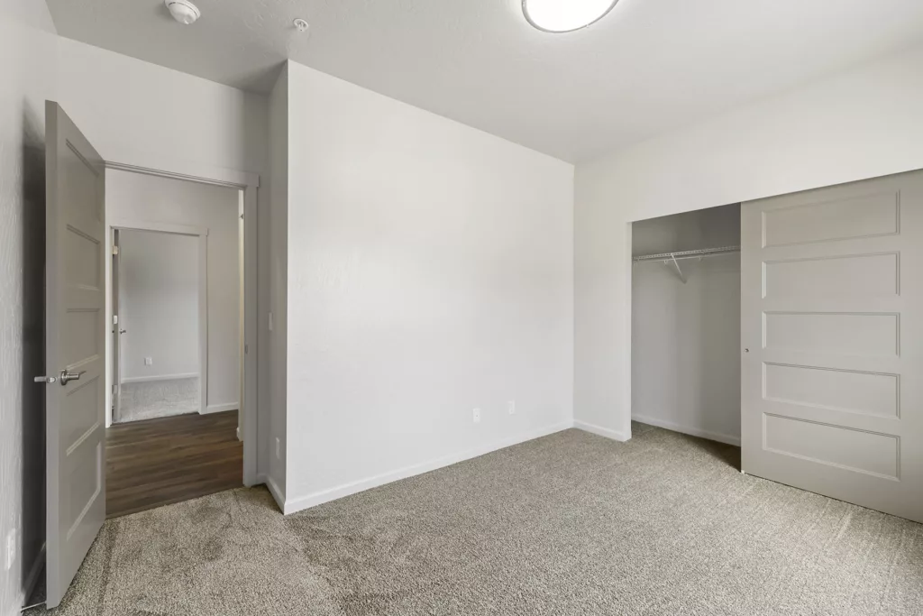 An empty room with light gray walls and carpeted floor, featuring an open closet with a sliding door and a door leading to another room with hardwood flooring.