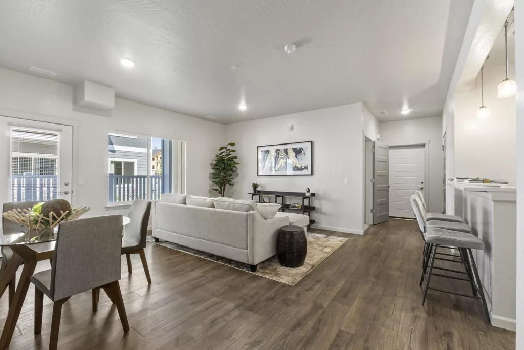 A modern living room with hardwood floors, featuring a light gray sofa, a glass dining table with gray chairs, and a bar area with stools. The room is well-lit with natural light from large windows and a glass door leading to a balcony. A framed artwork and a potted plant add to the decor.