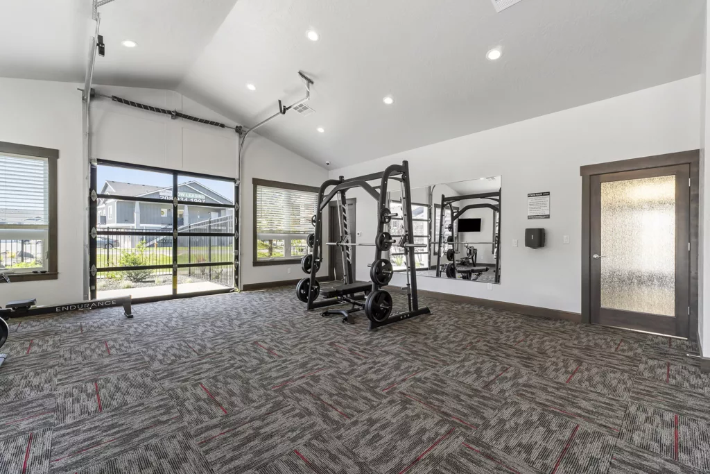 A modern gym with large windows, featuring a Smith machine with weights, a row machine labeled 'Endurance,' and a mirror on the wall. The room has carpeted flooring with a geometric pattern and is well-lit with recessed lighting.