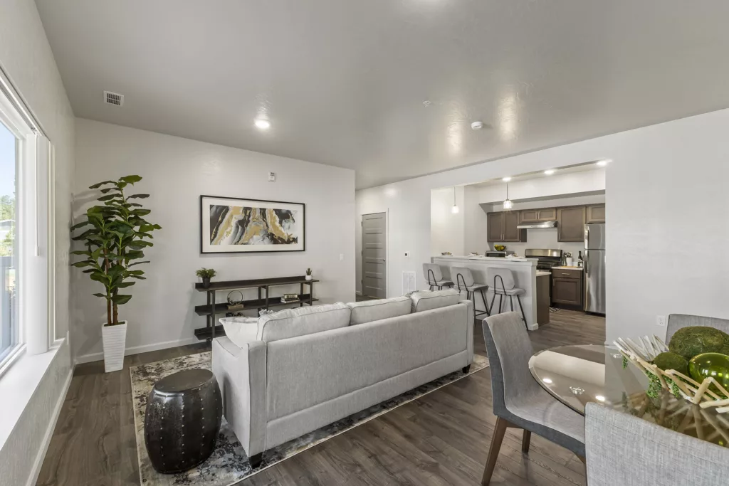 A modern living room with a light gray sofa, abstract wall art, and a potted plant, leading into a kitchen area with bar stools and stainless steel appliances.