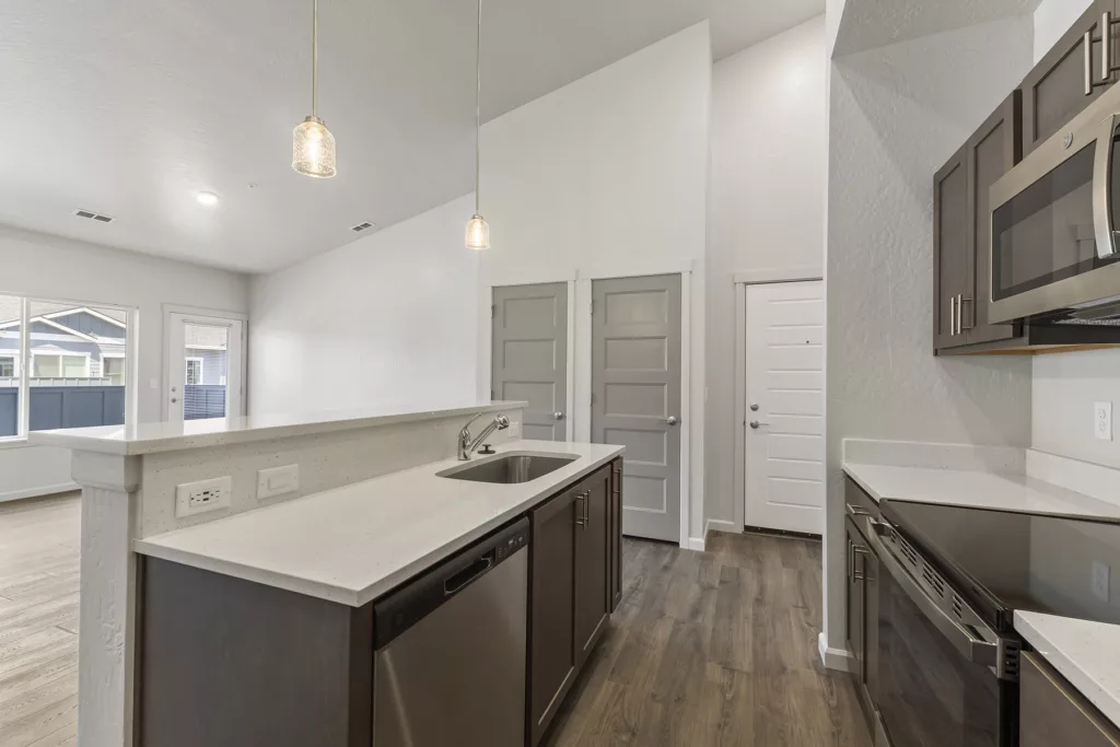 Modern kitchen interior with dark cabinetry, stainless steel appliances, white countertops, and pendant lighting, featuring a sink and dishwasher in the foreground and an open view to an adjacent room with a door and windows in the background.