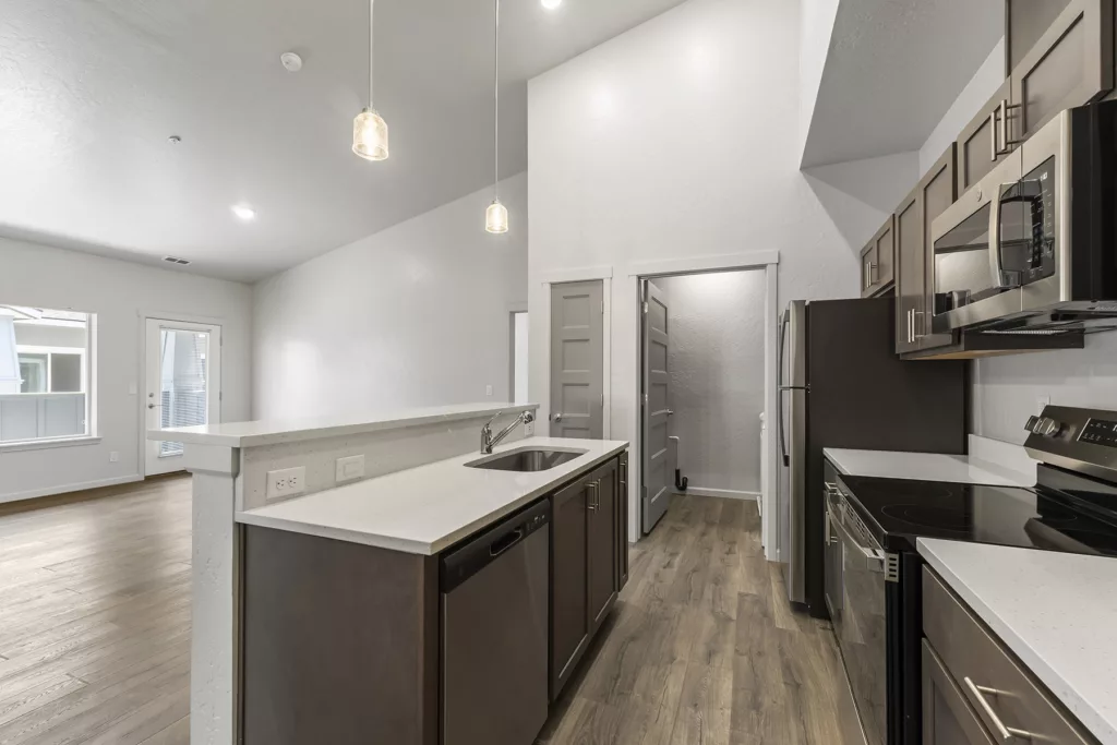 Modern kitchen with dark wood cabinets, stainless steel appliances, and a breakfast bar with pendant lighting. The kitchen opens into a living area with a glass door leading outside and wood-style flooring throughout.