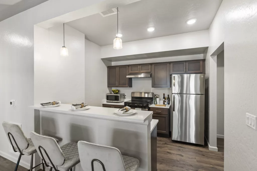 Modern kitchen with dark wooden cabinets, stainless steel appliances, and a white countertop with three padded stools and pendant lighting.