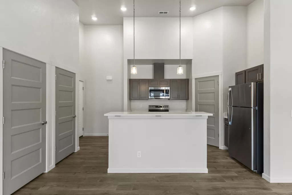 Modern kitchen with white countertops, stainless steel appliances, and dark wooden cabinets. The space is lit by pendant lights and features gray doors and wood flooring.