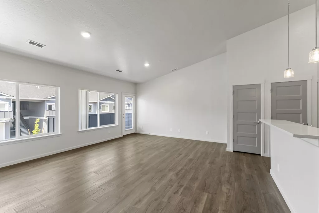 Spacious, empty room with light wood flooring, white walls, and large windows overlooking neighboring buildings. The room has a high ceiling with recessed lighting and pendant lights hanging above a corner counter next to two closed doors.