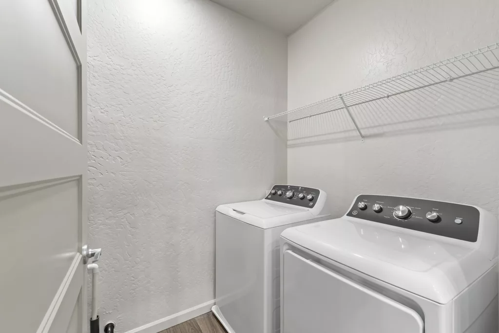 A laundry room with a white washing machine and dryer placed side by side, with a wire shelf above them attached to a textured white wall.