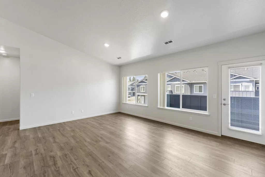 Empty living room with light wood flooring, two large windows, and a glass door leading outside, allowing natural light to enter.