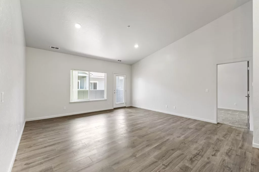 An empty living room with light wood flooring, white walls, a large window, and a glass door leading to a balcony. The ceiling is vaulted, and there is a doorway leading to another room with carpet flooring.