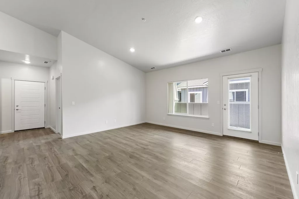 A spacious, empty room with white walls, wood flooring, and a high ceiling. It features a large window and a door with blinds, both allowing natural light to enter. The entrance door is visible on the left side of the room.