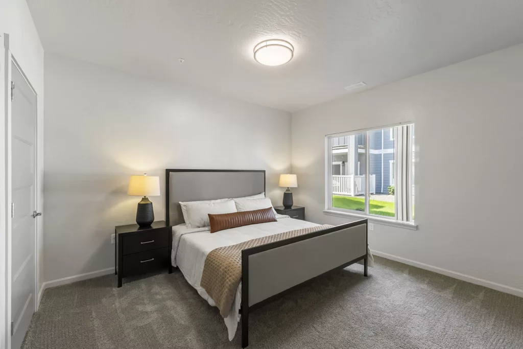A bedroom with a large bed featuring a gray upholstered headboard and footboard, two matching black nightstands with lamps, and a window overlooking a green lawn.