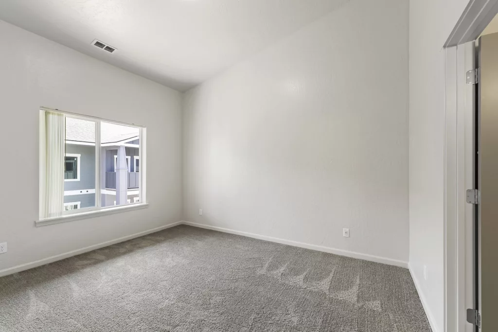 Empty room with light gray carpet, white walls, and a window showing a view of another building's exterior.