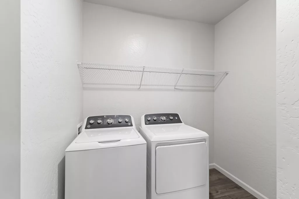 A laundry room with a white washing machine and dryer side by side beneath a wire shelf mounted on the wall.