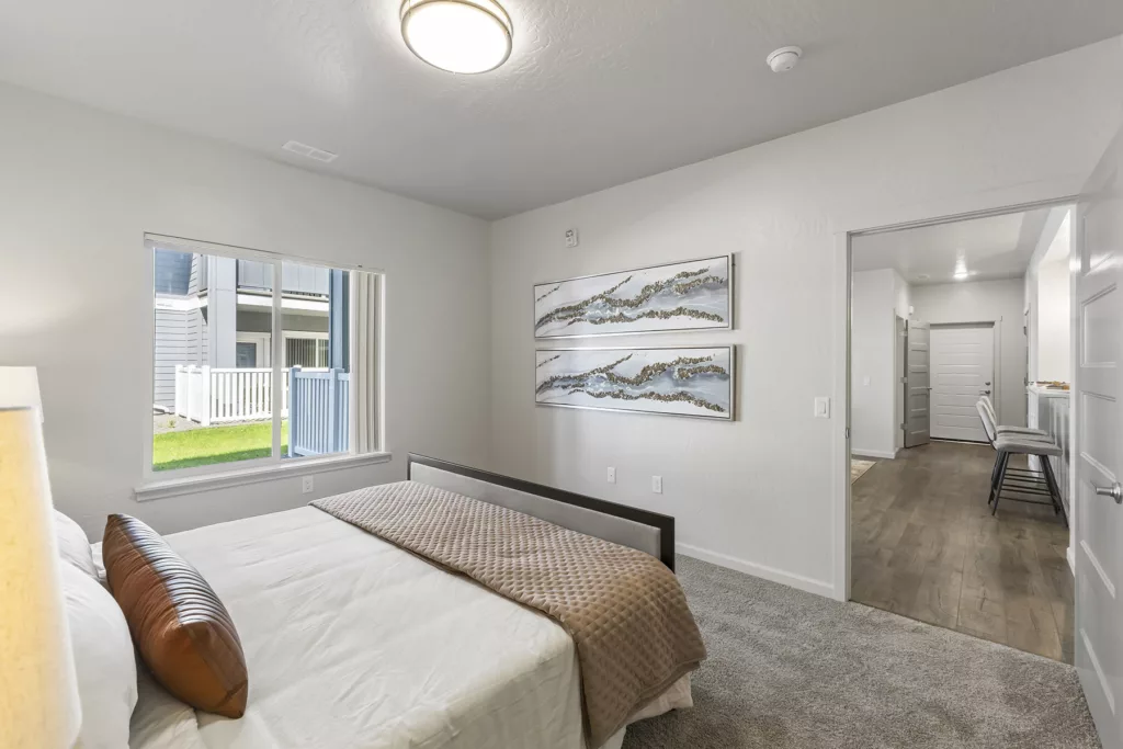A modern bedroom with a bed featuring white sheets and a beige quilt, a large window with a view of a grassy area, abstract art on the wall, and a doorway leading to a hallway with bar stools.