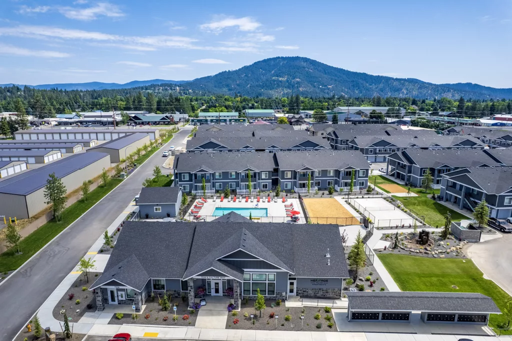 Aerial view of a residential complex featuring multiple gray two-story buildings surrounding a central swimming pool area with red lounge chairs. In the background, a wooded mountain range is visible under a clear blue sky.