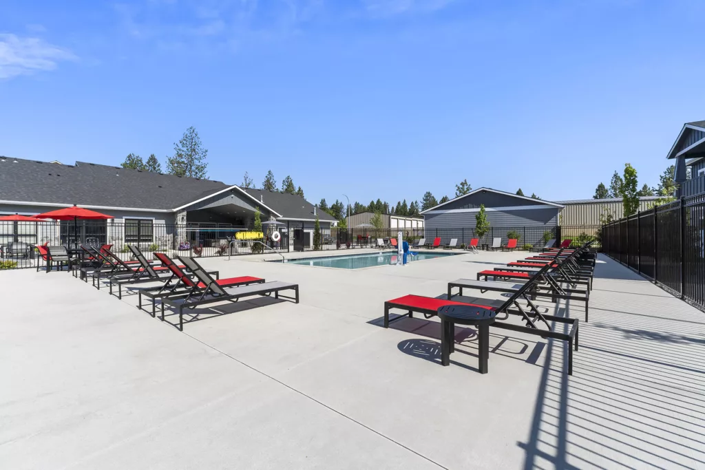 Outdoor swimming pool area with multiple red-cushioned lounge chairs, tables, and umbrellas on a sunny day. The pool is surrounded by fencing and is adjacent to a building with a dark roof.