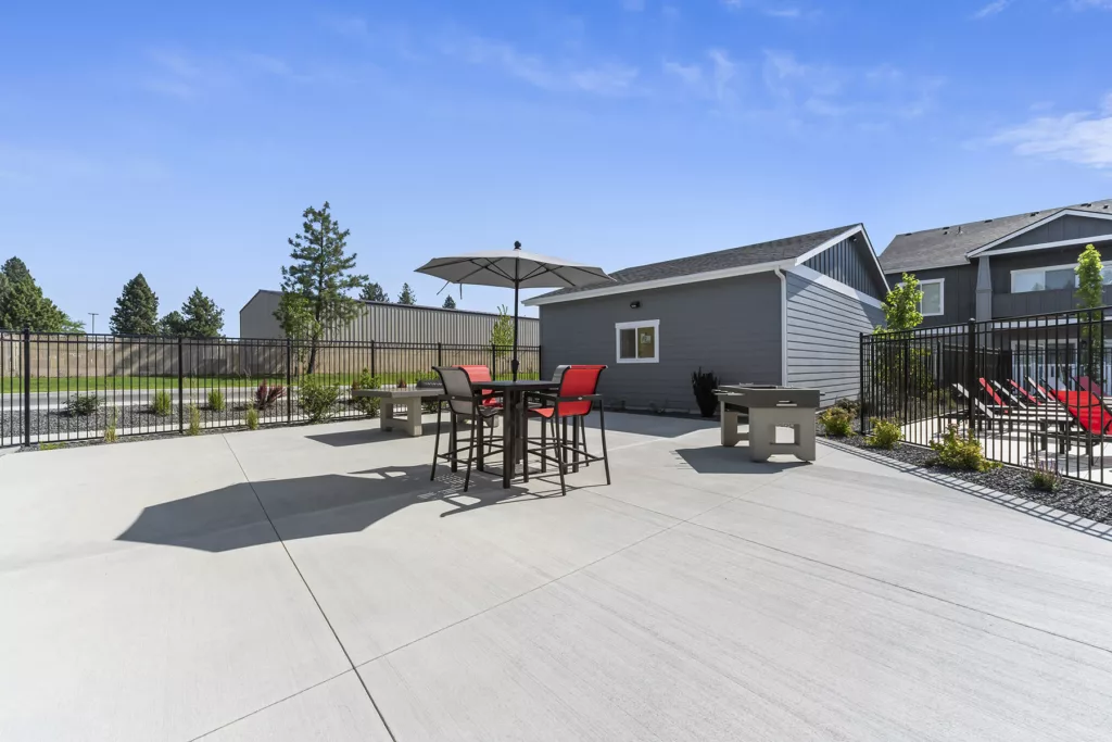 Outdoor patio area with a table, red chairs, and an umbrella, surrounded by a fence and adjacent to a gray building under a clear blue sky.