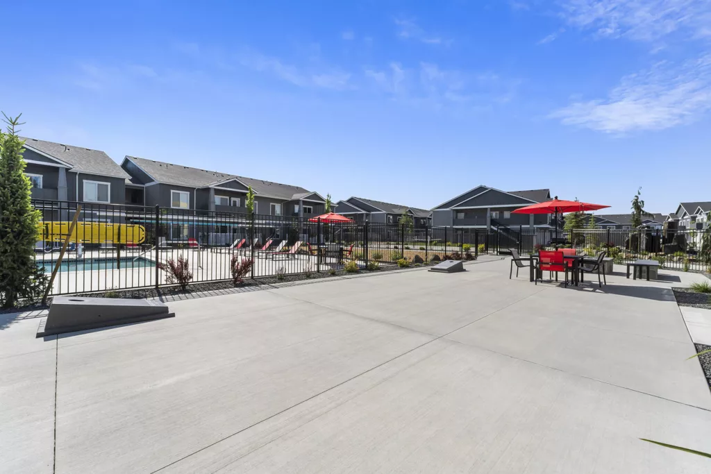 Outdoor area with a concrete patio, featuring a table with red chairs and a red umbrella. A fence surrounds a swimming pool area with more red umbrellas and lounge chairs, while townhouse-style buildings are visible in the background under a clear blue sky.