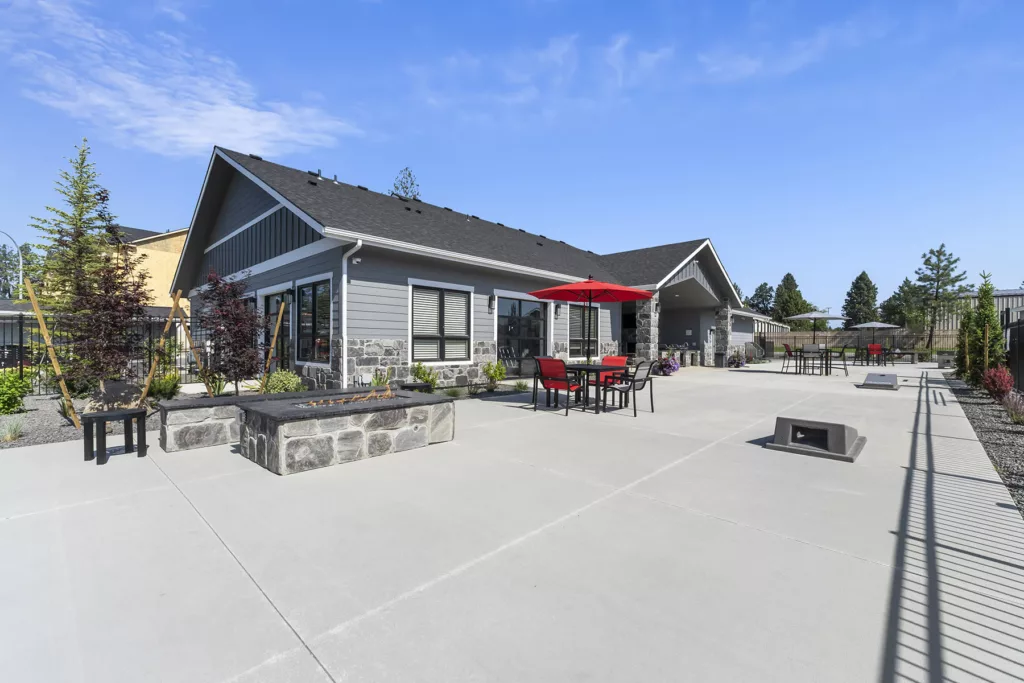 A patio area with outdoor seating and tables, featuring red umbrellas and chairs, adjacent to a modern building with stone accents.