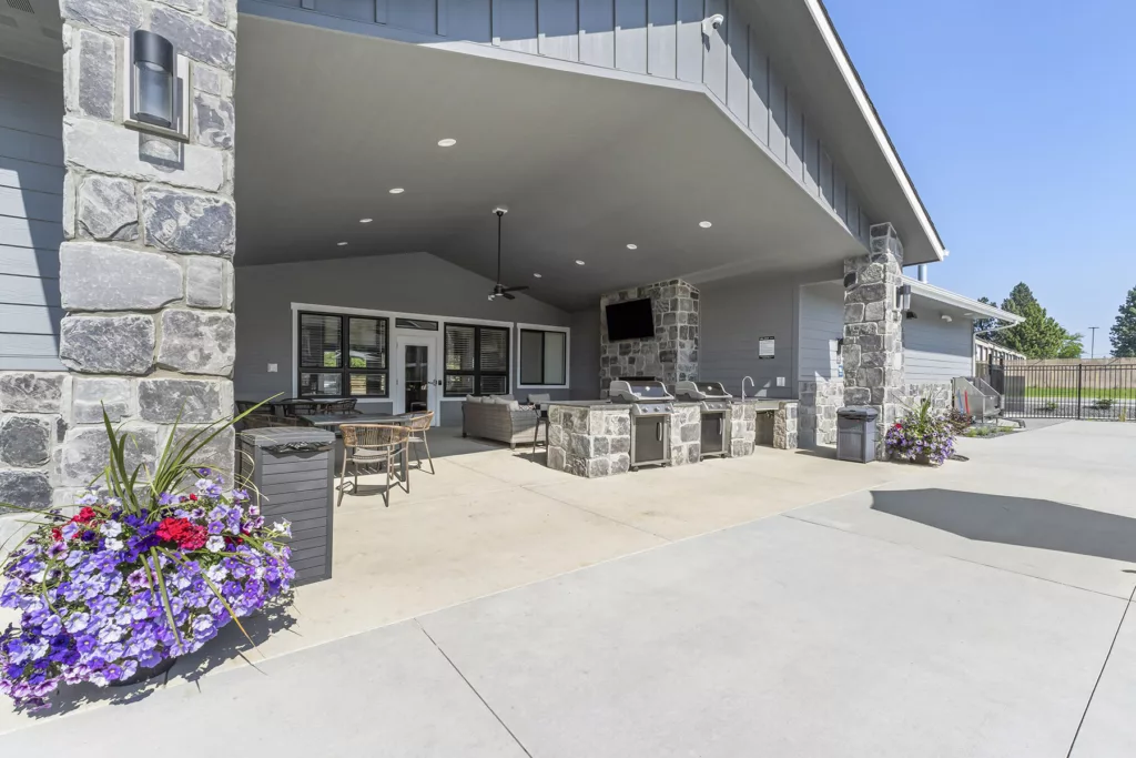 Outdoor patio area with stone columns, seating, barbecue grills, and a mounted TV, surrounded by vibrant flower arrangements.