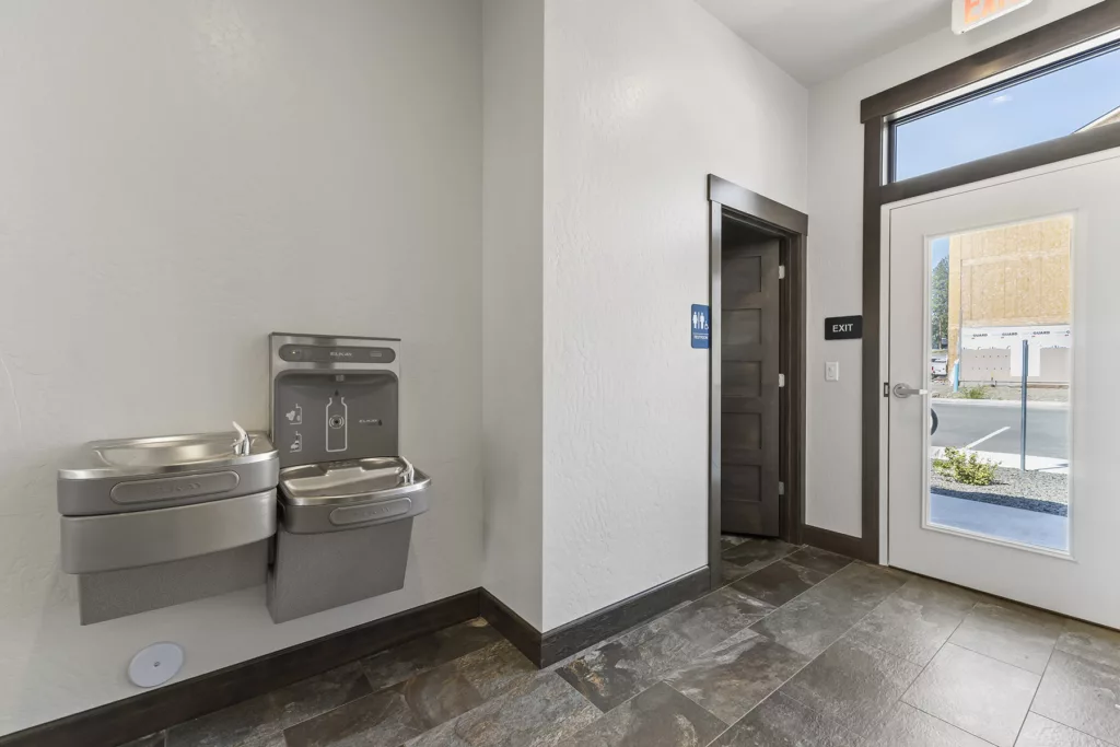 A room with a water fountain and bottle refill station next to a restroom door and an exit leading outside.