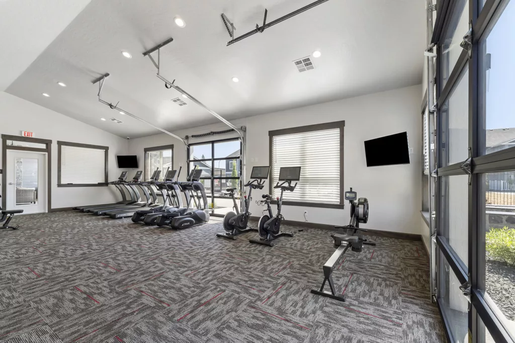 A modern gym with several treadmills, stationary bikes, and a rowing machine on a carpeted floor. The room features high ceilings, large windows, and a flat-screen TV mounted on the wall.