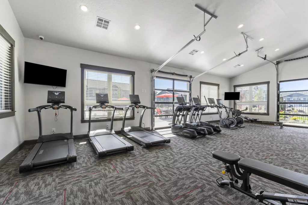 A spacious, well-lit gym featuring several treadmills, elliptical machines, and a bench press, with large windows allowing natural light to illuminate the workout area.